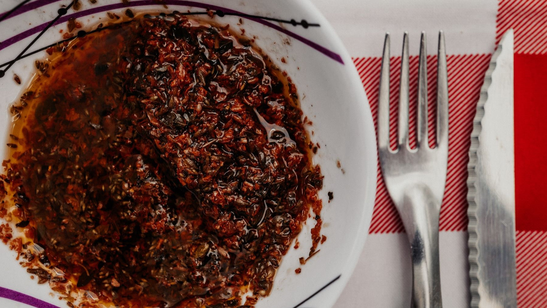 A rustic bowl of herb-rich chimichurri sits beside cutlery on a checked tablecloth.