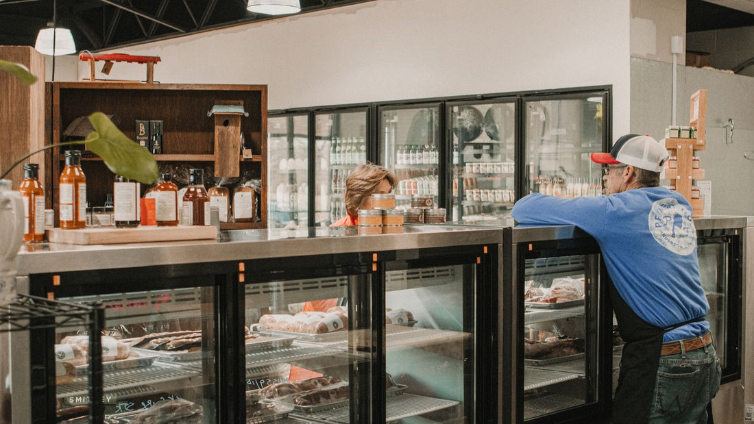 A customer speaks with a butcher across refrigerated display cases filled with fresh cuts.