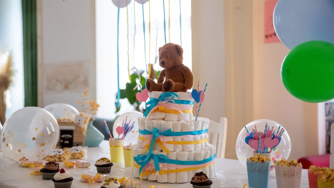A baby shower table is decorated with balloons, cupcakes, a teddy bear and a colourful nappy cake.