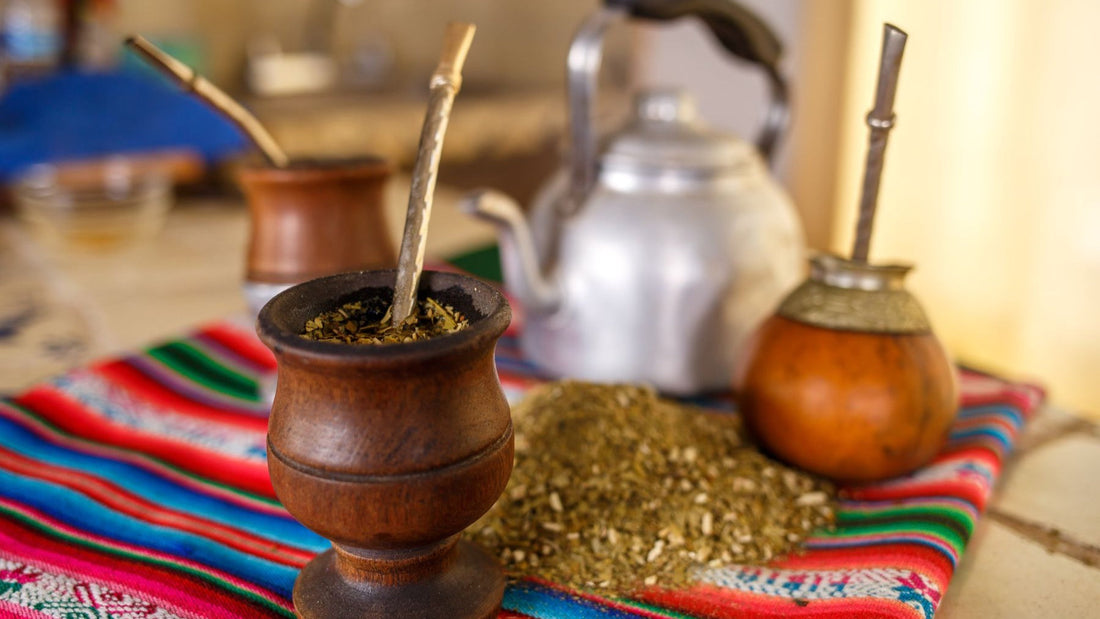Several traditional mate gourds, bombillas and loose yerba are arranged on a colourful woven cloth.