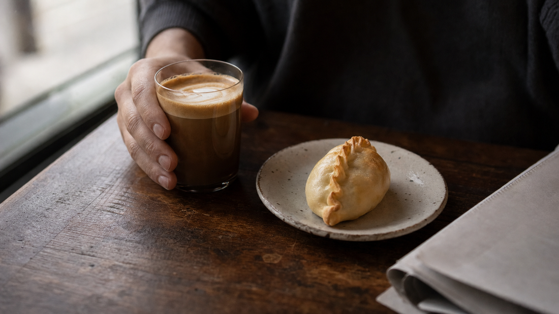 A hand lifts a glass of coffee beside one sealed golden empanada on a ceramic plate at a quiet Sydney café table.