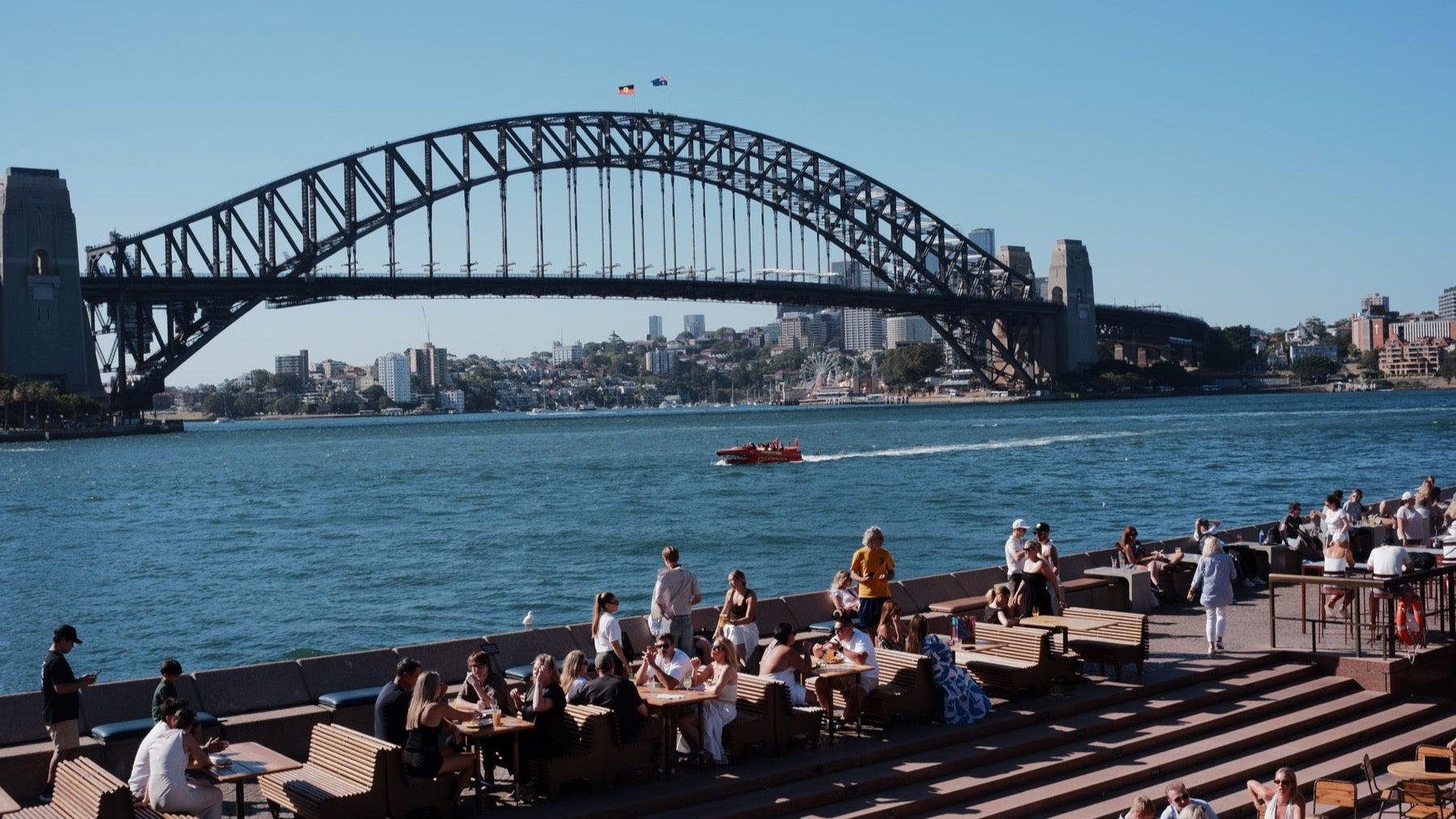 People dine outdoors beside Sydney Harbour with the Harbour Bridge spanning the background.