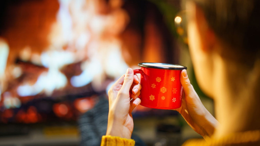 A person holds a warm drink in front of a glowing fireplace during a cosy winter night at home.