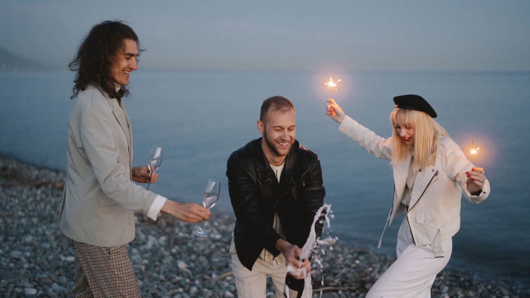 Friends celebrate beside the ocean at twilight with champagne and sparklers.