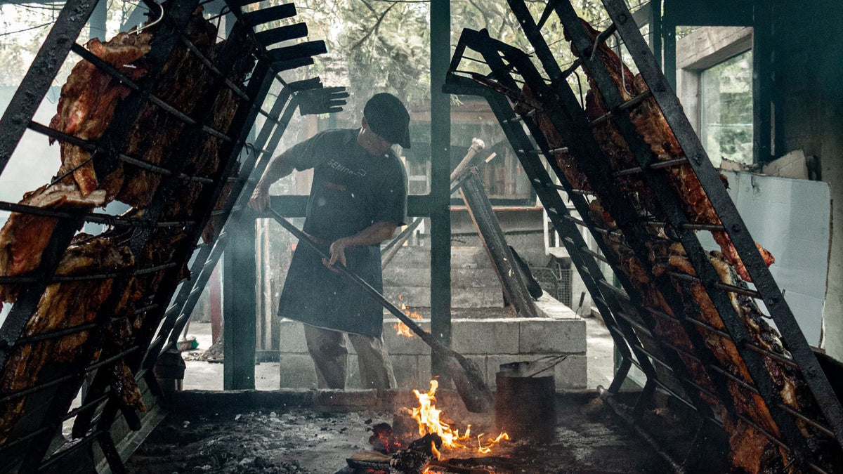 An asador tends a central wood fire between upright racks of meat in a traditional Argentine barbecue.