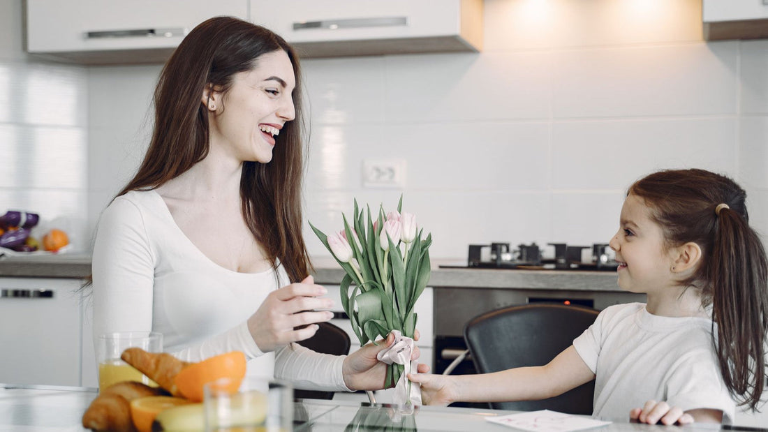 A smiling daughter gives her mother a bouquet of tulips over breakfast in the kitchen.