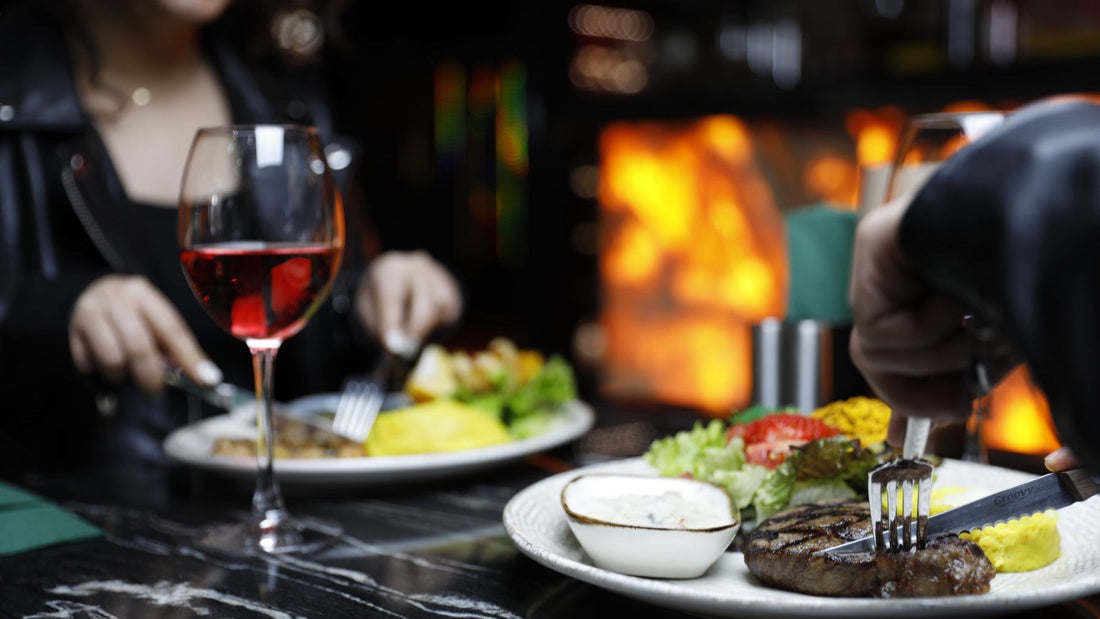 Diners enjoy grilled steak and red wine beside an open fire in a warmly lit restaurant.