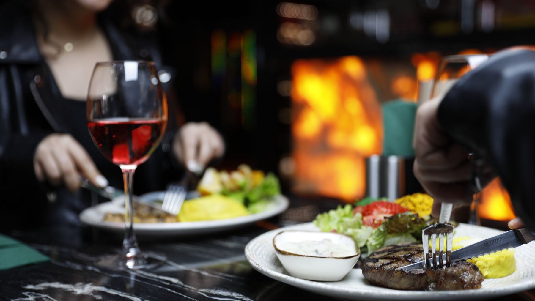 Diners enjoy grilled steak and red wine beside an open fire in a warmly lit restaurant.