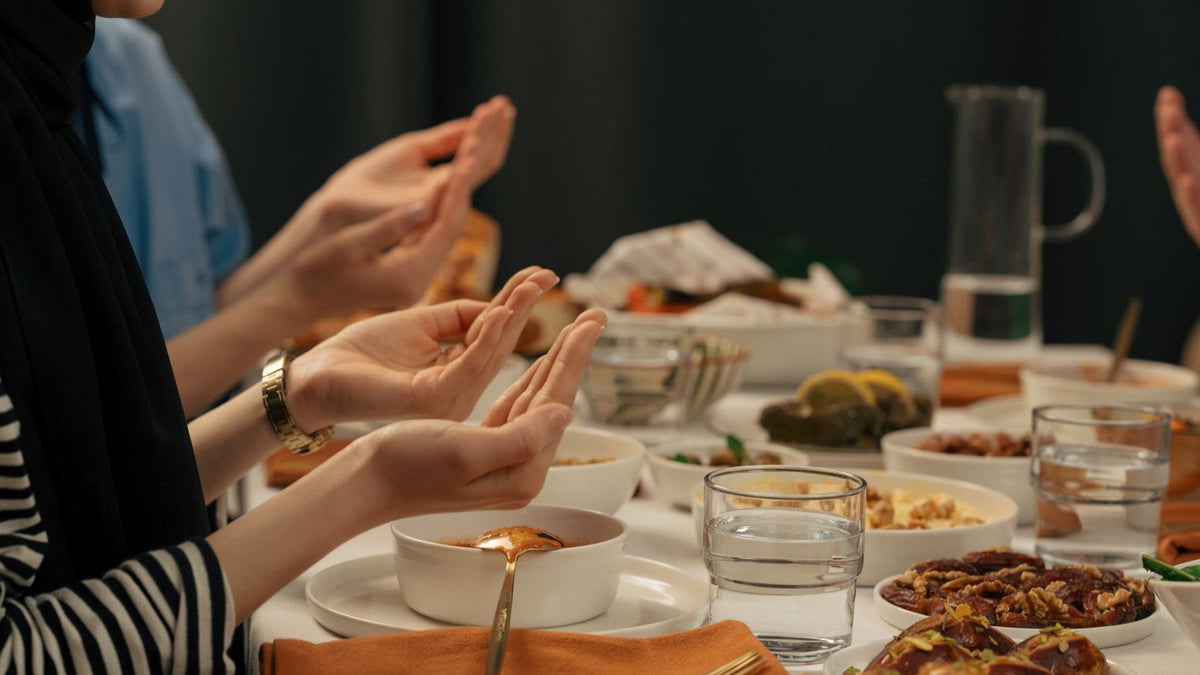 Hands are raised in prayer around a Ramadan iftar table set with dates, soup and shared dishes.