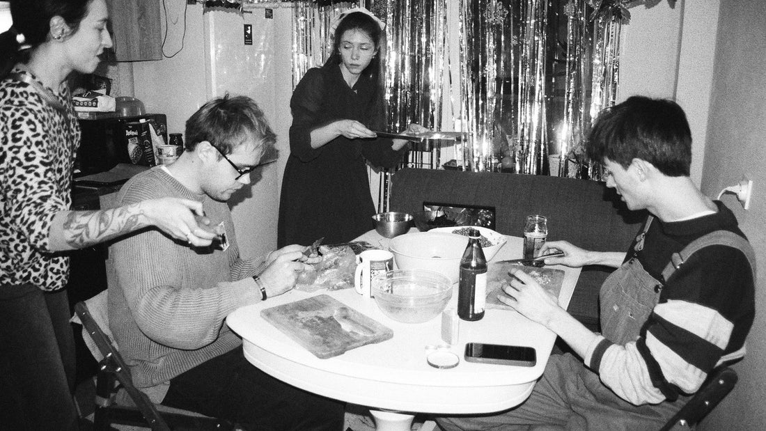 Friends prepare and share food around a compact dining table inside a small apartment.
