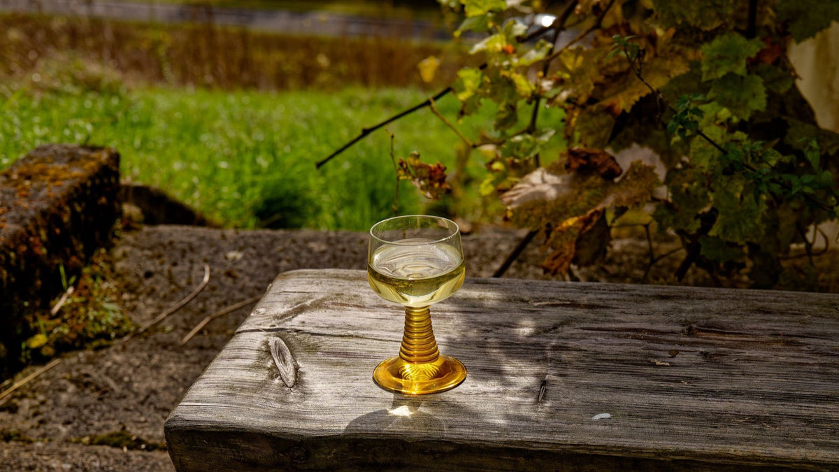 A glass of pale white wine rests on a rustic table beside vineyard vines.