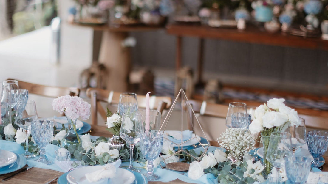 An outdoor wedding reception table is arranged with blue place settings, glassware, flowers and greenery.