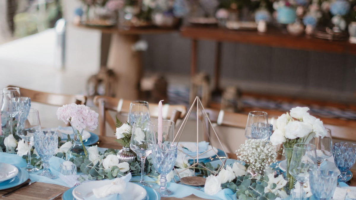 An outdoor wedding reception table is arranged with blue place settings, glassware, flowers and greenery.