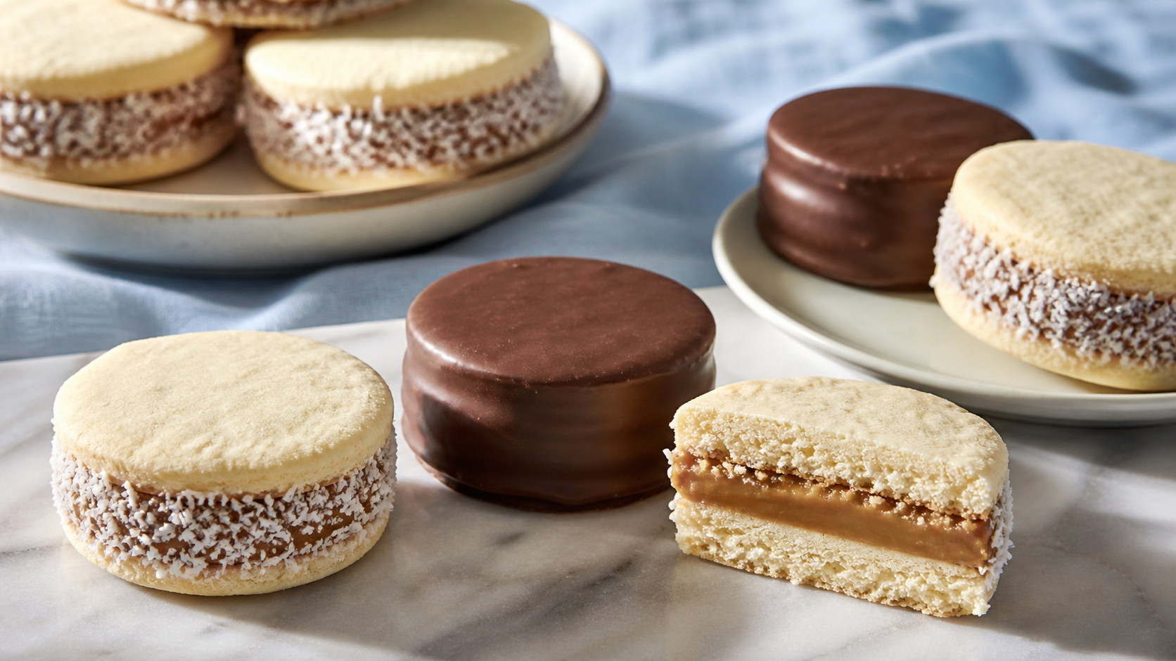 Classic alfajores with dulce de leche, coconut and chocolate coatings arranged on marble.
