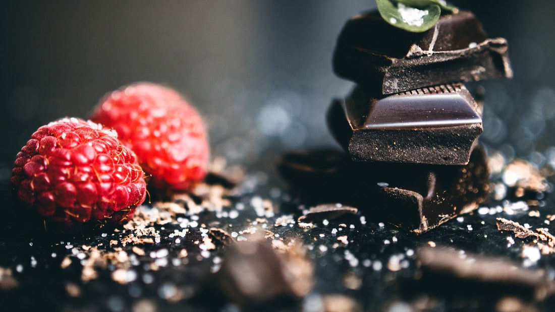 Fresh raspberries sit beside pieces of dark chocolate on a dramatic black surface.
