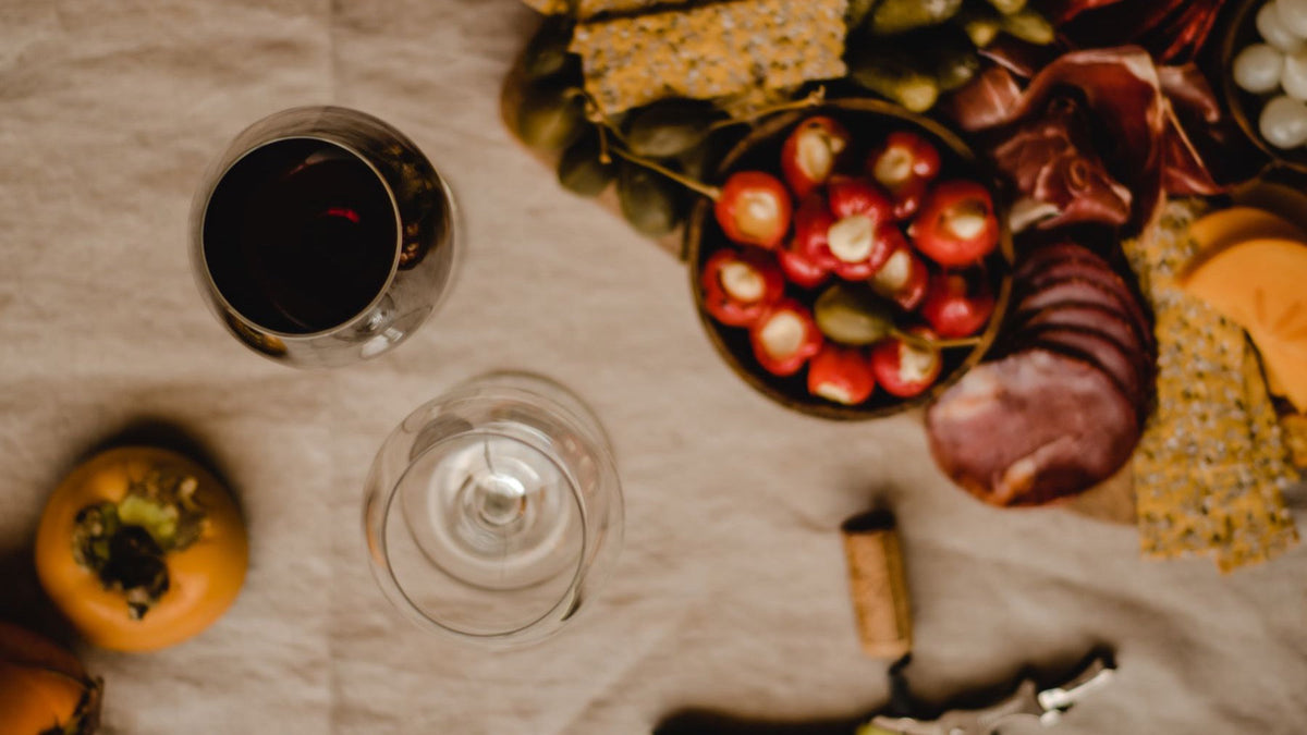A glass of red wine sits beside a grazing board of cured meats, crackers and pickled vegetables.