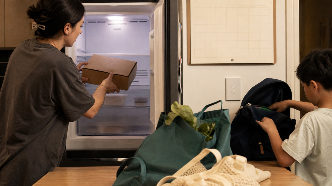 A parent stores a closed kraft freezer box while a child packs a school bag beside reusable groceries and a blank family calendar.