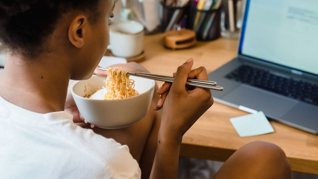 A woman eats noodles beside her laptop during a work-from-home lunch break.