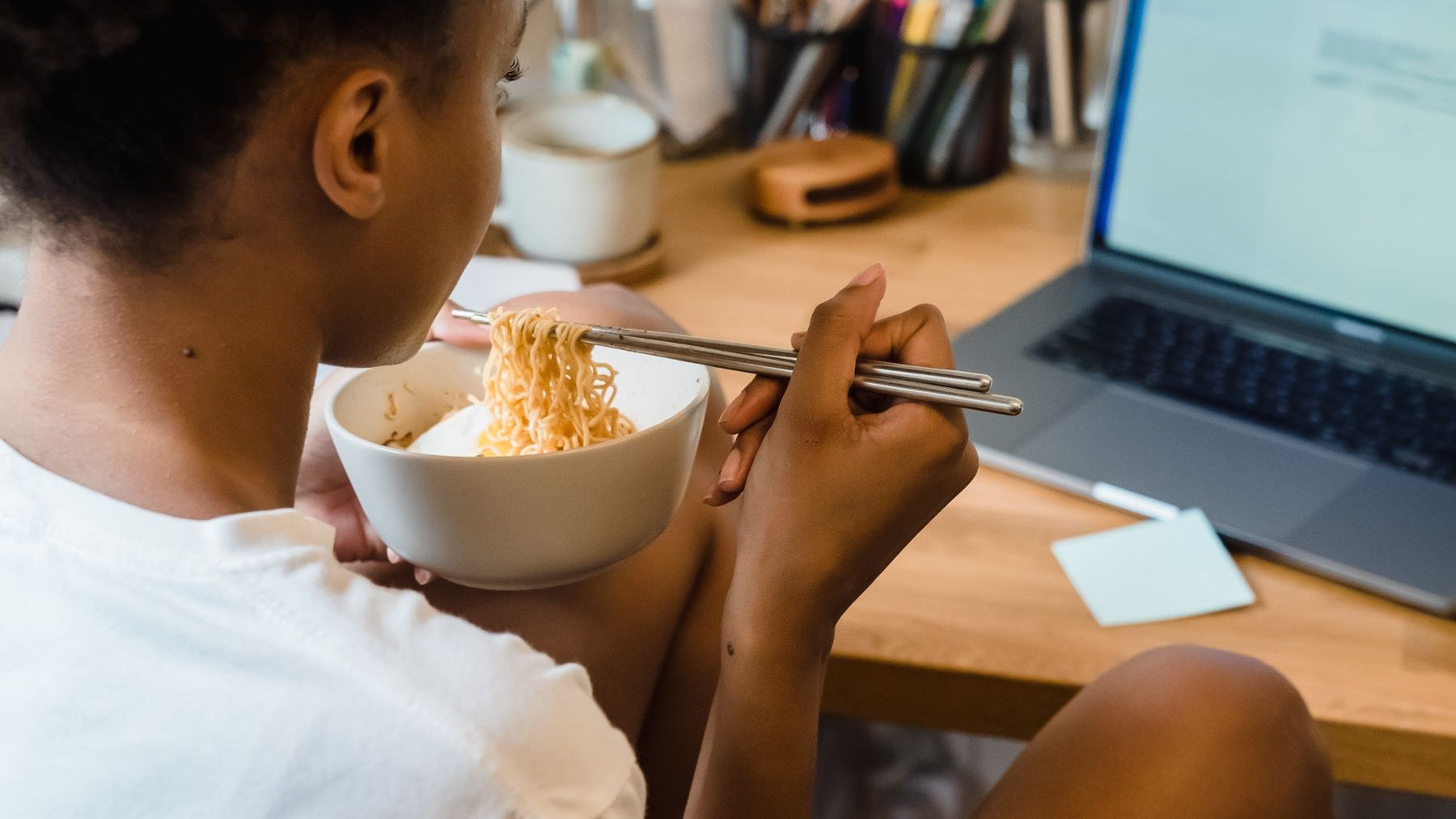 A woman eats noodles beside her laptop during a work-from-home lunch break.