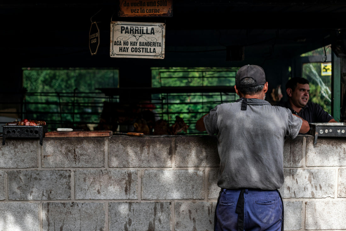 A man ordering at a traditional Argentine parrilla grill counter with a handwritten sign reading Acá no hay banderita, hay costilla