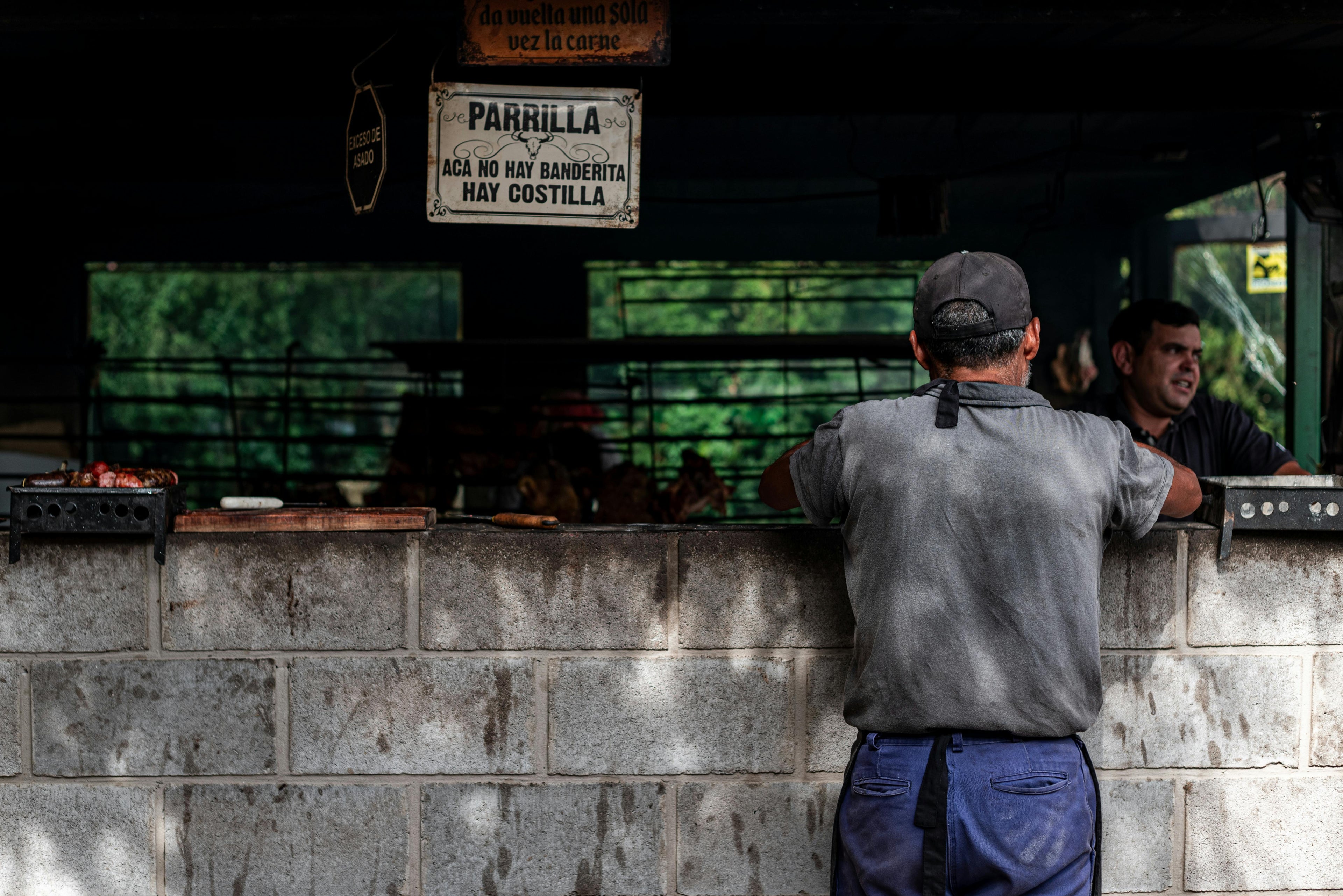 A man ordering at a traditional Argentine parrilla grill counter with a handwritten sign reading Acá no hay banderita, hay costilla