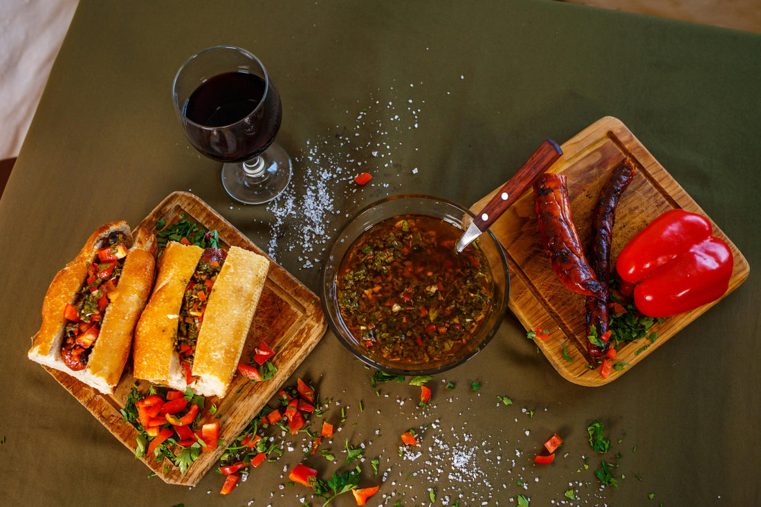 Overhead view of an Argentinian spread featuring a choripán sandwich on a wooden board, a bowl of chimichurri sauce, grilled chorizo with red capsicum, and a glass of red wine on a dark green table