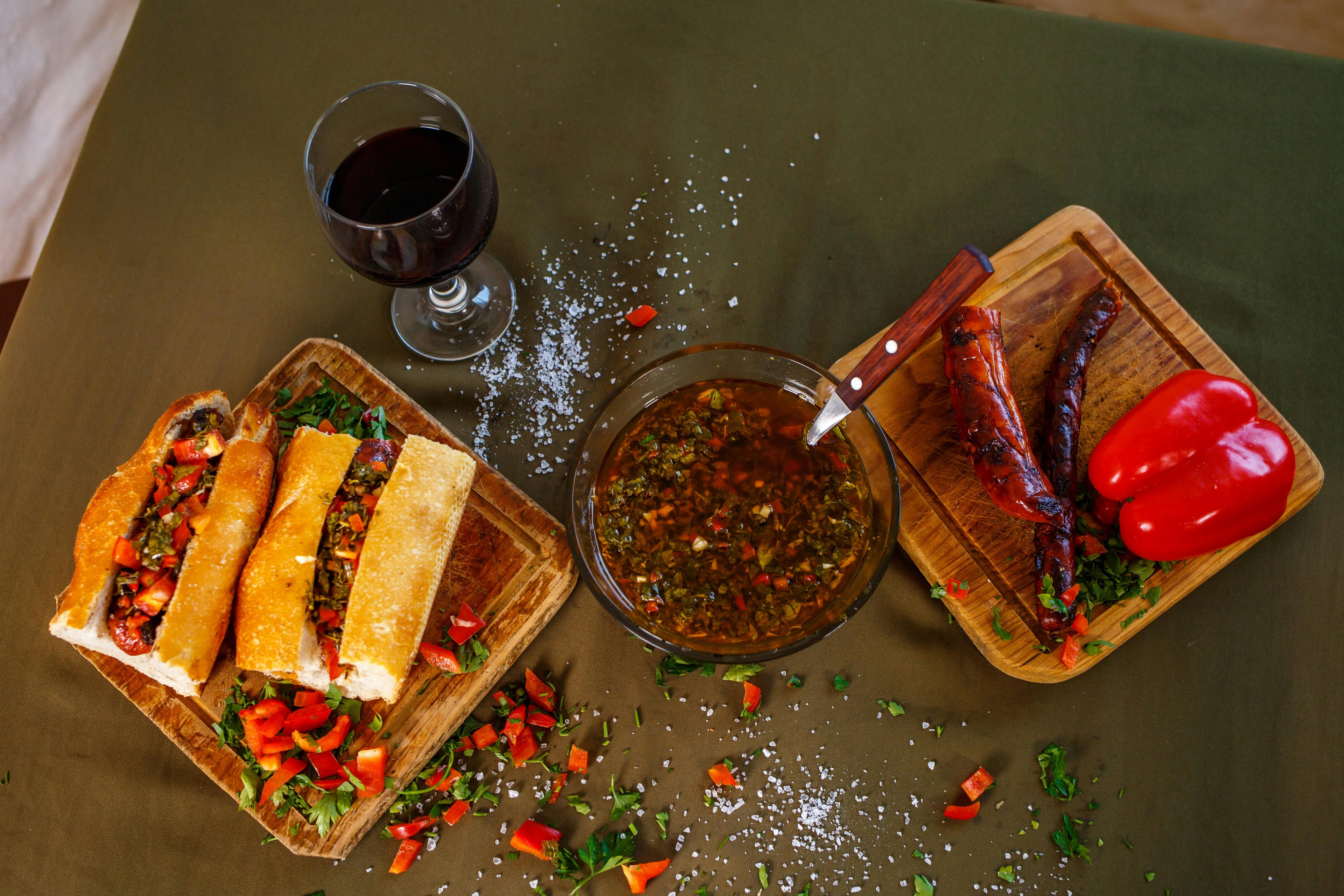 Overhead view of an Argentinian spread featuring a choripán sandwich on a wooden board, a bowl of chimichurri sauce, grilled chorizo with red capsicum, and a glass of red wine on a dark green table