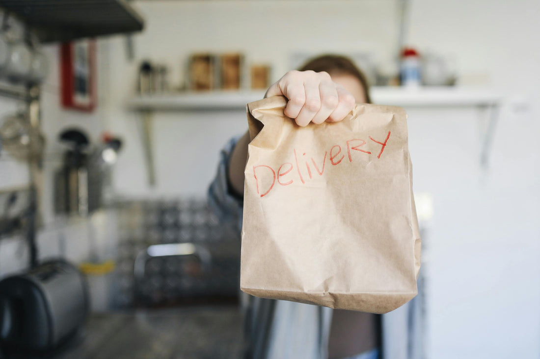 Person holding a brown paper delivery bag with the word Delivery written in red, in a kitchen setting
